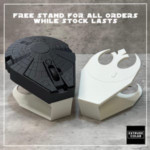 May include: A black computer mouse shaped like a circular spacecraft with intricate surface detailing, resting on a white plastic stand. Beside it is an identical white stand featuring a cutout of a stylized rebel alliance symbol emblem.
