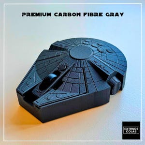 May include: A dark gray, textured, 3D-printed model of a spaceship with a circular main body and a forward-projecting cockpit. The text "PREMIUM CARBON FIBRE GRAY" is printed above the object, and a logo for "EXTRUDE COLAB" is in the corner.
