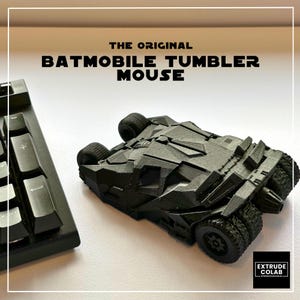 May include: A black computer mouse shaped like the Batmobile Tumbler vehicle. The top surface features the text THE ORIGINAL BATMOBILE TUMBLER MOUSE. A black keyboard is visible on the left side, and an EXTRUDE COLAB logo is in the corner.