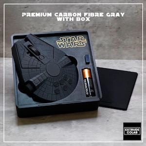 May include: A matte gray computer mouse shaped like the Millennium Falcon from Star Wars, housed in a custom black tray with a Duracell battery. The text PREMIUM CARBON FIBRE GRAY WITH BOX is visible at the top, and the Star Wars logo is printed in gold.