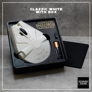 May include: A white computer mouse shaped like the Millennium Falcon from Star Wars, housed in a black rectangular box with a gold Star Wars logo. The set includes a black Duracell battery and a small black component, all arranged in a custom tray.
