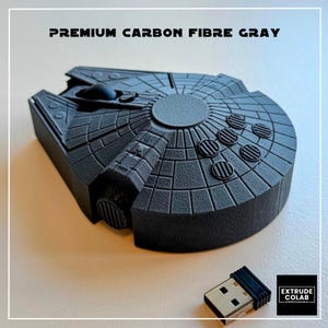 May include: A dark gray computer mouse shaped like a spaceship with a textured surface, labeled PREMIUM CARBON FIBER GRAY. The device features a circular central section and a separate USB receiver with a metallic connector and black plastic casing.