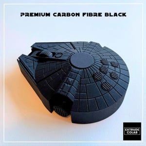May include: A matte black computer mouse shaped like a spaceship with intricate circular and grid-like surface detailing. The text "PREMIUM CARBON FIBRE BLACK" is printed at the top, and a small logo box for "EXTRUDE COLAB" is in the bottom corner.
