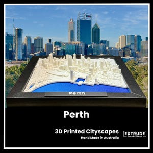 May include: A 3D printed cityscape model of Perth, Australia. The model is white and shows the city's skyline, including the Swan River. The model is framed in black and sits on a blue base. The text "Perth" is printed on the base.