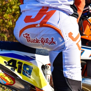 May include: A person wearing white and orange motocross pants with a "Chick-fil-A" logo on the back. The person is sitting on a white and blue dirt bike with a yellow number "418" on the side.