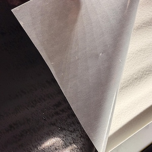 May include: A sheet of adhesive transfer paper with a translucent, matte finish being peeled back to reveal a smooth, off-white backing layer. The top layer is semi-transparent, showing a subtle texture, while the bottom layer is solid and opaque.