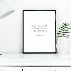 May include: A framed print with a black border displays the quote "We did not come to this earth to gain our worth; we brought it with us" by Ardeth G. Kapp. The print is on a white surface, next to a white vase with a green plant.