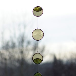 May include: A hanging stained glass suncatcher with five circular pieces. The glass is a mix of clear and olive green, with a crescent moon design on some. The suncatcher is suspended by a thin wire, creating a delicate, decorative piece.