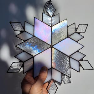 May include: A decorative stained glass snowflake with a black metal frame. The snowflake features clear, white, and iridescent glass pieces in a geometric design. The glass reflects light, creating a shimmering effect.