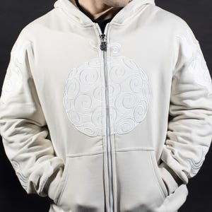 Puede incluir: Sudadera con capucha con cremallera de color crema con un diseño bordado circular grande en la parte delantera. La sudadera con capucha tiene una capucha, bolsillos y detalles intrincados en las mangas. La cremallera tiene un tirador decorativo. La persona lleva pantalones negros.