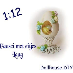 Könnte beinhalten: Ein cremefarbenes, eiförmiges Ornament mit einem Ausschnitt, der Miniatur-Eier in Stroh zeigt. Das Ornament ist mit pastellfarbenen Blumen verziert und steht auf einem detaillierten Sockel. Text enthält "1:12" und "Dollhouse DIY".
