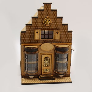 Puede incluir: Un modelo de madera de una casa de estilo holandés con una fachada de ladrillo, una puerta decorativa y dos ventanas grandes. La casa está sobre una pequeña base de madera con un patrón de ladrillo.