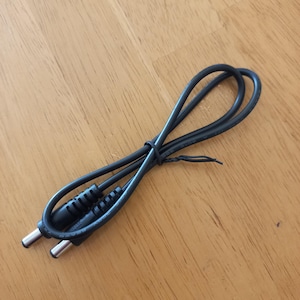 Op de afbeelding: Een zwart stroomsnoer met twee stekkers aan de uiteinden. Het snoer is opgerold en met een zwart elastiekje vastgebonden.