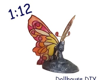 Op de afbeelding: Een vlinderfiguur op schaal 1:12 met doorschijnende vleugels in roze, oranje en gele tinten, voorzien van donkere omlijningen. De vlinder is gemonteerd op een grijze, getextureerde basis. De tekst 1:12 en Dollhouse DIY zijn zichtbaar.