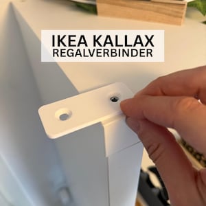 May include: A white IKEA KALLAX REGALVERBINDER shelf connector is held near a white shelf corner. The connector has a rectangular shape with a hole and a screw. The text "IKEA KALLAX REGALVERBINDER" is displayed in a white rectangle.