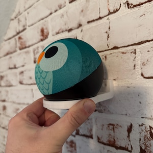 Op de afbeelding: Een witte wandbeugel voor een slimme speaker met een uil-ontwerp. De speaker is teal en zwart met een wit en oranje oog.