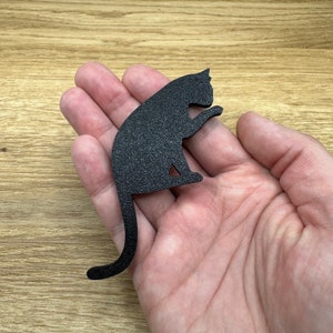 May include: Black cat silhouette cutout, made of a textured material, sitting on a hand.