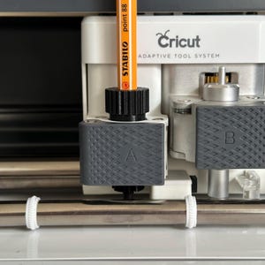 Könnte beinhalten: Nahaufnahme einer Cricut-Maschine mit einem Bleistift, der in den Werkzeughalter eingesetzt ist. Der Werkzeughalter ist mit "A" gekennzeichnet und die Maschine trägt den Schriftzug "Cricut Adaptive Tool System".