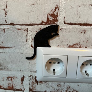 May include: A black cat silhouette wall decor is mounted on a white brick wall next to a white electrical outlet with two plugs.