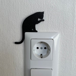 May include: A black cat silhouette sticker is placed on a white electrical outlet cover. The outlet cover has a standard electrical outlet and a light switch.