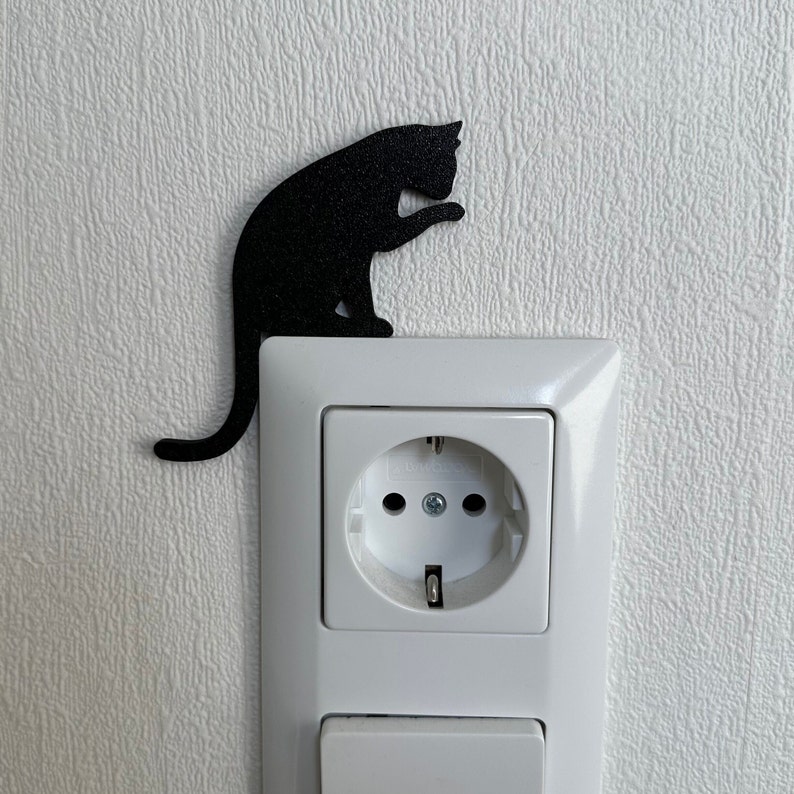 May include: A black cat silhouette sticker is placed on a white electrical outlet cover. The outlet cover has a standard electrical outlet and a light switch.
