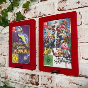 Può includere: Due custodie per giochi Nintendo Switch rosse con coperchi in plastica trasparente. La custodia a sinistra contiene una cartuccia di gioco Pokemon Purple. La custodia a destra contiene una cartuccia di gioco Super Smash Bros. Ultimate.