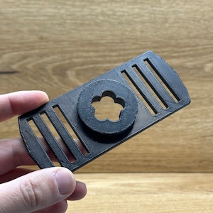 Op de afbeelding: Een zwart, 3D-geprint object van plastic met een bloemvormige uitsparing en vijf parallelle lijnen. Het object is ontworpen om te worden gebruikt als telefoonstandaard.