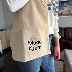 Könnte beinhalten: Eine beigefarbene Tragetasche mit dem Text "Finger weg Muddi Kram" in Schwarz. Die Tasche besteht aus einem hellfarbenen Stoff. Die Tasche wird von einer Person getragen, die einen weißen Pullover und eine blaue Jeans trägt.