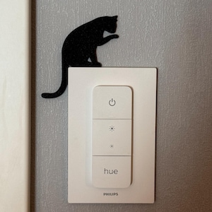 May include: A white Philips Hue smart light switch with a black cat silhouette sticker on the top. The switch has three buttons: on/off, brightness up, and brightness down. The switch also has the word "hue" and the Philips logo.