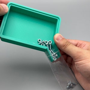 Op de afbeelding: Een turquoise rechthoekige bak met een deksel wordt in de handen gehouden. Kleine zilveren bouten en moeren worden uit een doorzichtige plastic zak in de bak gegoten. De bak heeft een glad oppervlak en een opstaande rand.
