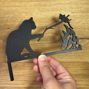 Könnte beinhalten: Schwarze Metallsilhouette einer Katze, die einen Stock mit einem Vogel am Ende hält, der über einem Lagerfeuer brät. Die Silhouette ist auf einem Metallständer montiert.