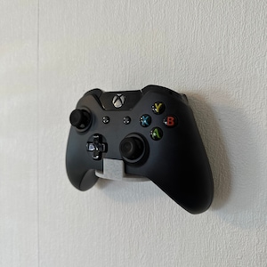 May include: A black Xbox controller with white buttons and a gray wall mount. The controller has a black and white Xbox logo on the front.