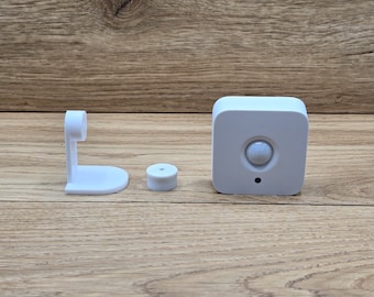 May include: A white motion sensor with a round sensor lens, a white mounting bracket, and a white base. The sensor is on a wooden surface.