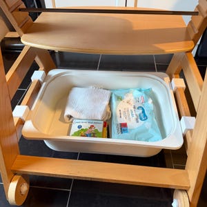Könnte beinhalten: Ein hölzerner Hochstuhl mit einem Tablett und einem unteren Regal mit einer weißen Kunststoffwanne. Die Wanne enthält ein gefaltetes weißes Tuch, ein Buch und eine Packung Babytücher. Der Stuhl hat Räder.