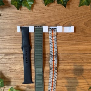 Op de afbeelding: Drie horlogebandjes hangen aan een witte, wandgemonteerde organizer. De bandjes zijn zwart, groen en zilver met oranje accenten.