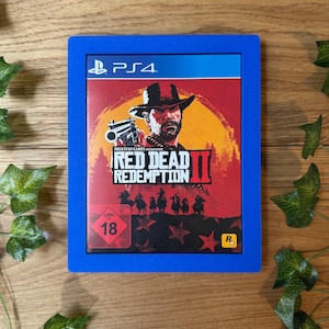 Könnte beinhalten: Ein blaues PlayStation 4-Spielgehäuse mit einem roten, weißen und schwarzen Cover, das den Titel "Red Dead Redemption II" und einen Cowboy-Charakter mit einer Waffe zeigt. Das Spiel ist von der ESRB mit "R" bewertet und hat eine USK-Bewertung von 18.