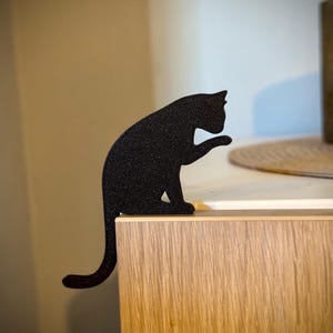 May include: Black cat silhouette figurine sitting on a wooden surface. The cat is in a sitting position with its tail curled around its body.