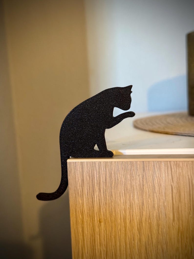 May include: Black cat silhouette figurine sitting on a wooden surface. The cat is in a sitting position with its tail curled around its body.