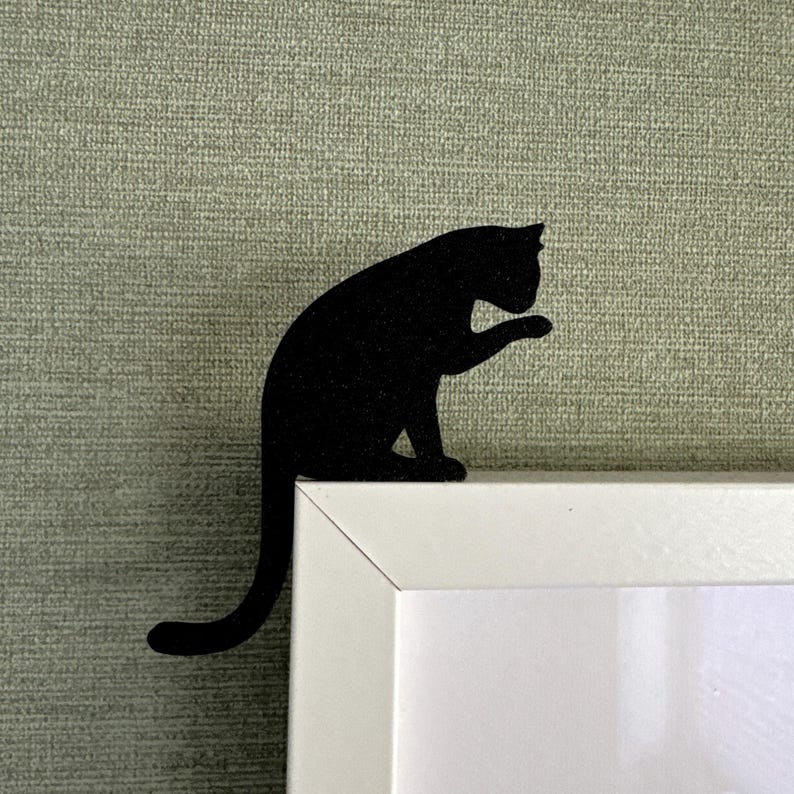 May include: Black cat silhouette bookmark, sitting on a white frame. The cat's tail is curled around the frame.