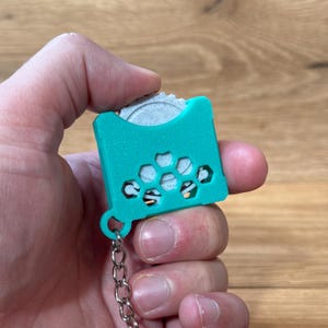 May include: A teal blue keychain with a small, round, white disc inside. The keychain has a honeycomb pattern and a silver chain.