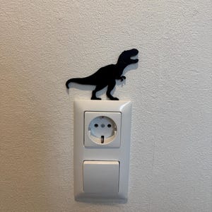 Könnte beinhalten: Eine schwarze Dinosauriersilhouette ist über einer weißen Steckdose und einem Lichtschalter montiert.