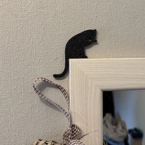 May include: A black cat silhouette wall decor with a tail that hangs down. The cat is sitting on a white wooden frame with a mirror.