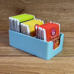 Op de afbeelding: Een lichtblauwe theezakjeshouder met vier vakken. De houder is gevuld met theezakjes in verschillende smaken, waaronder "Mediterraan" en "Italiaans".