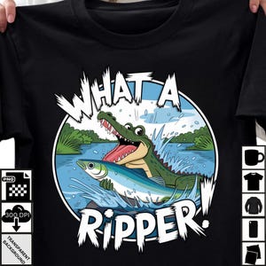 Peut inclure: T-shirt noir avec un dessin animé d'un alligator vert attrapant un poisson bleu dans l'eau. Le texte "WHAT A RIPPER!" est en blanc. Le design comprend des éclaboussures d'eau et des plantes vertes.
