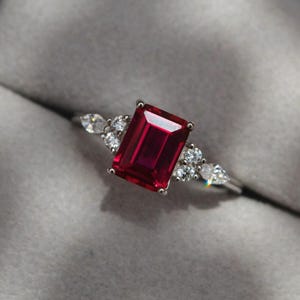 Puede incluir: Un anillo de plata con una gran piedra de rubí de talla esmeralda rodeada de diamantes redondos más pequeños.