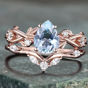 May include: A rose gold engagement ring set featuring a pear-shaped, light blue gemstone. The ring is accented with small, round, and marquise-cut white gemstones. The band has a floral design.