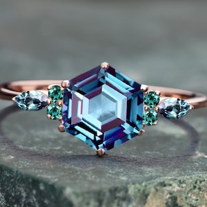 Könnte beinhalten: Ein roségoldener Ring mit einem großen, sechseckigen, blau-grünen Edelstein im Mittelpunkt. Kleinere teal- und grüne Edelsteine flankieren den zentralen Stein. Der Ring liegt auf einer strukturierten, grün-grauen Oberfläche.