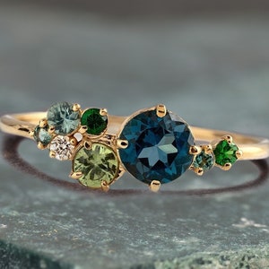 Op de afbeelding: Een gouden ring met een cluster van blauwe, groene en witte edelstenen. De ring heeft een grote blauwe edelsteen in het midden, omringd door kleinere groene en witte edelstenen.