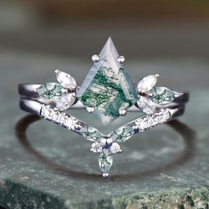 May include: A silver engagement ring set featuring a kite-shaped moss agate center stone with green inclusions. The ring is flanked by smaller white gemstones and a curved band with additional small, clear stones. The ring sits on a green stone surface.