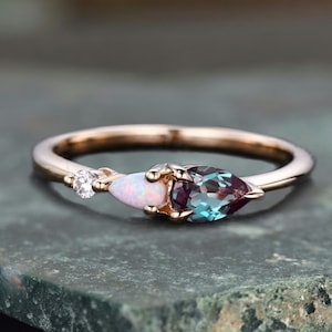 May include: A delicate rose gold ring with three pear-shaped gemstones. The center stone is a vibrant blue-green alexandrite, flanked by a white opal and a smaller clear diamond.
